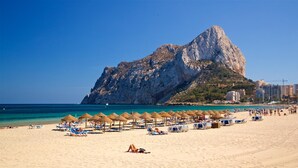 Ein Strand mit Liegestühlen und Sonnenschirmen, eine große Felsformation im Hintergrund und ein klarer, blauer Himmel.