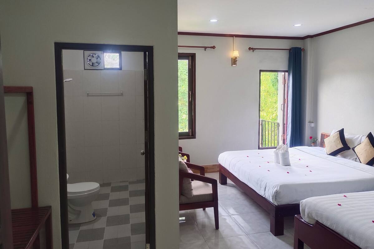 Ein Hotelzimmer mit Schachbrettmuster-Boden, einem Bett mit weißer Bettwäsche und dekorativen Kissen, einem Nachttisch und einem Fenster mit Blick auf Grünflächen.