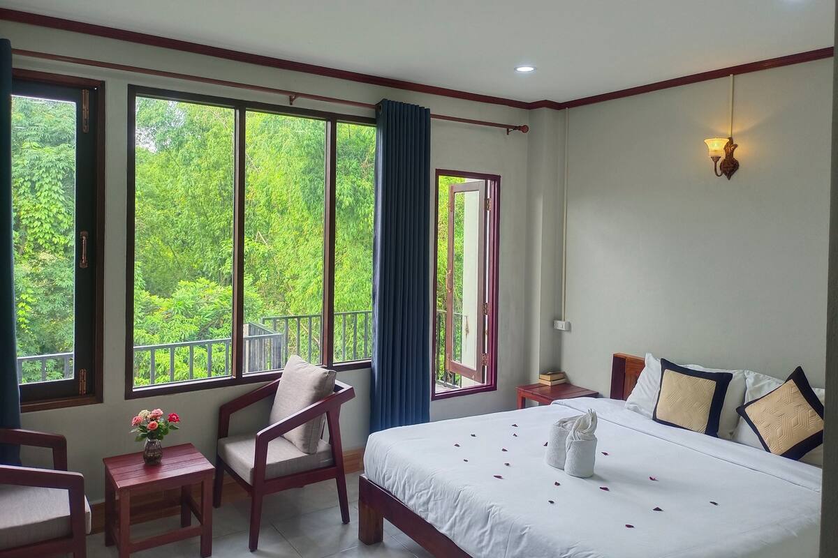 Ein Hotelzimmer mit einem großen Bett, einem Holzstuhl, einem kleinen Tisch mit Blumenvase und Blick auf üppiges Grün durch die Fenster.