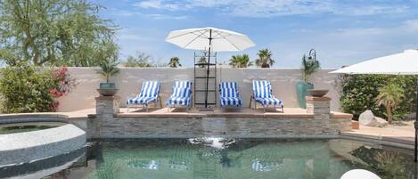 A pool area with blue and white striped lounge chairs, a white umbrella, and a hot tub.