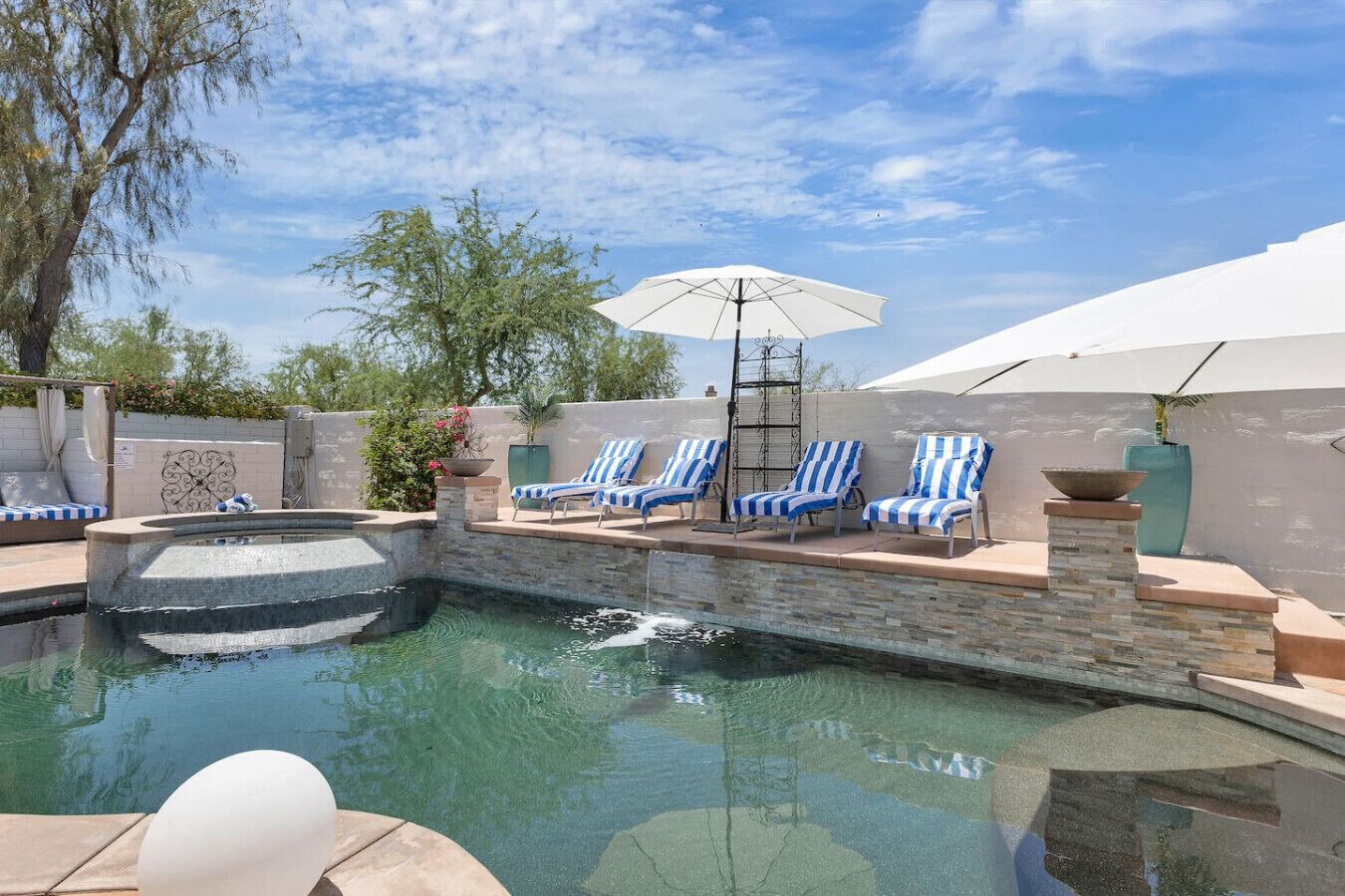 A swimming pool with blue and white striped lounge chairs, a white umbrella, and a stone wall.