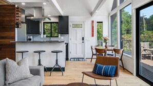 A modern kitchen with dark cabinets, a built-in oven, and a large window. There is a dining area with a table and chairs, and a sofa with patterned cushions. - Renovated A-frame with water view (Vashon)
