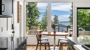 A modern kitchen with a large island, a dining area with a round table and four chairs, and a view of a deck and trees. - Renovated A-frame with water view (Vashon)
