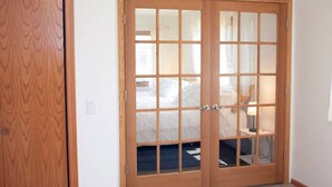 Puerta doble de madera con paneles de vidrio, que da acceso a un dormitorio con cama y mesita de noche.