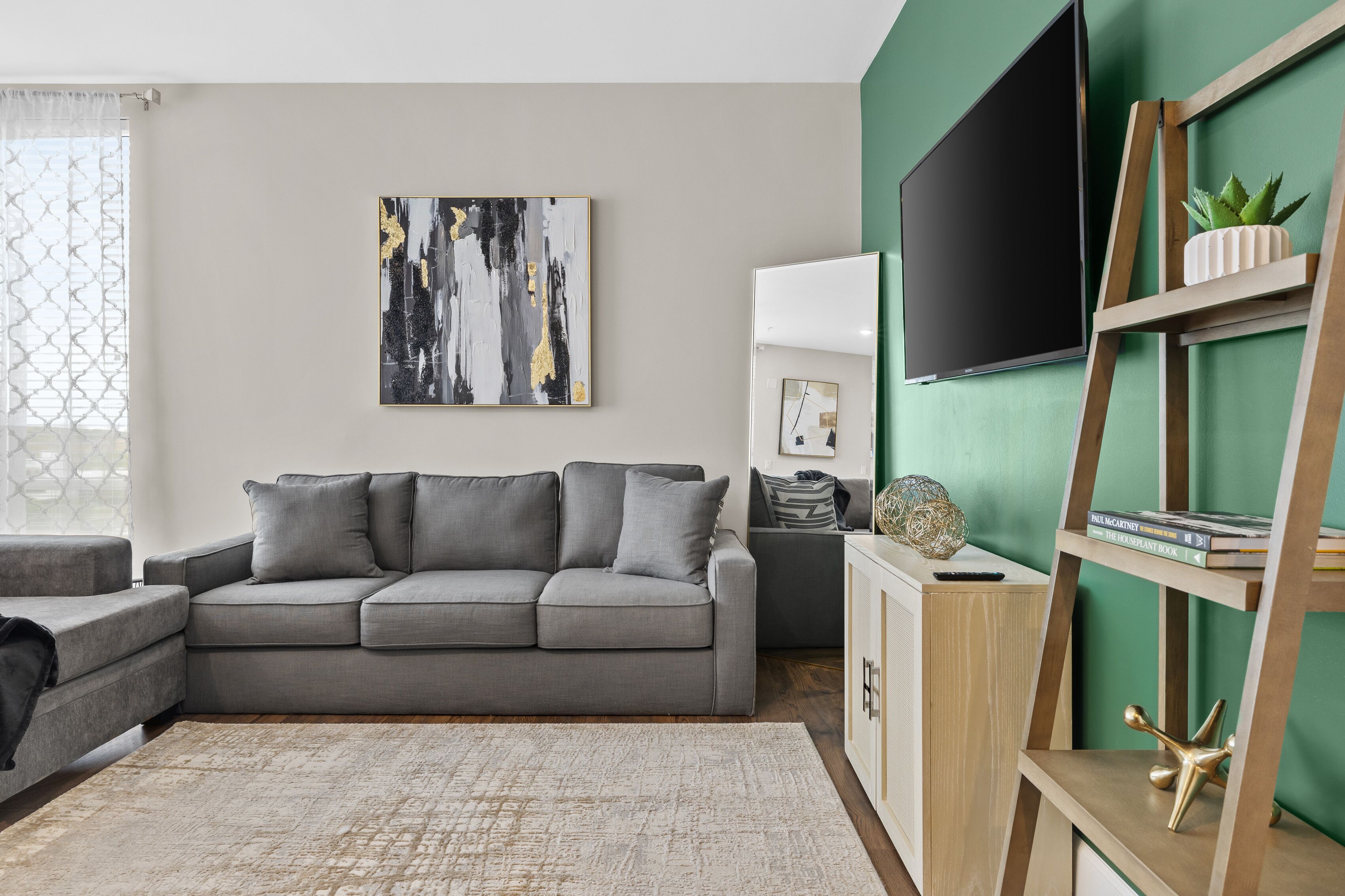 A modern living room with a grey sectional sofa, a large abstract painting, a flat-screen TV, a wooden ladder shelf, and a green accent wall.