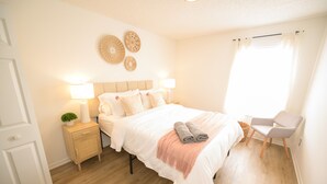 A neatly arranged bedroom with a bed, bedside lamps, a small nightstand, a chair, and a window with curtains.