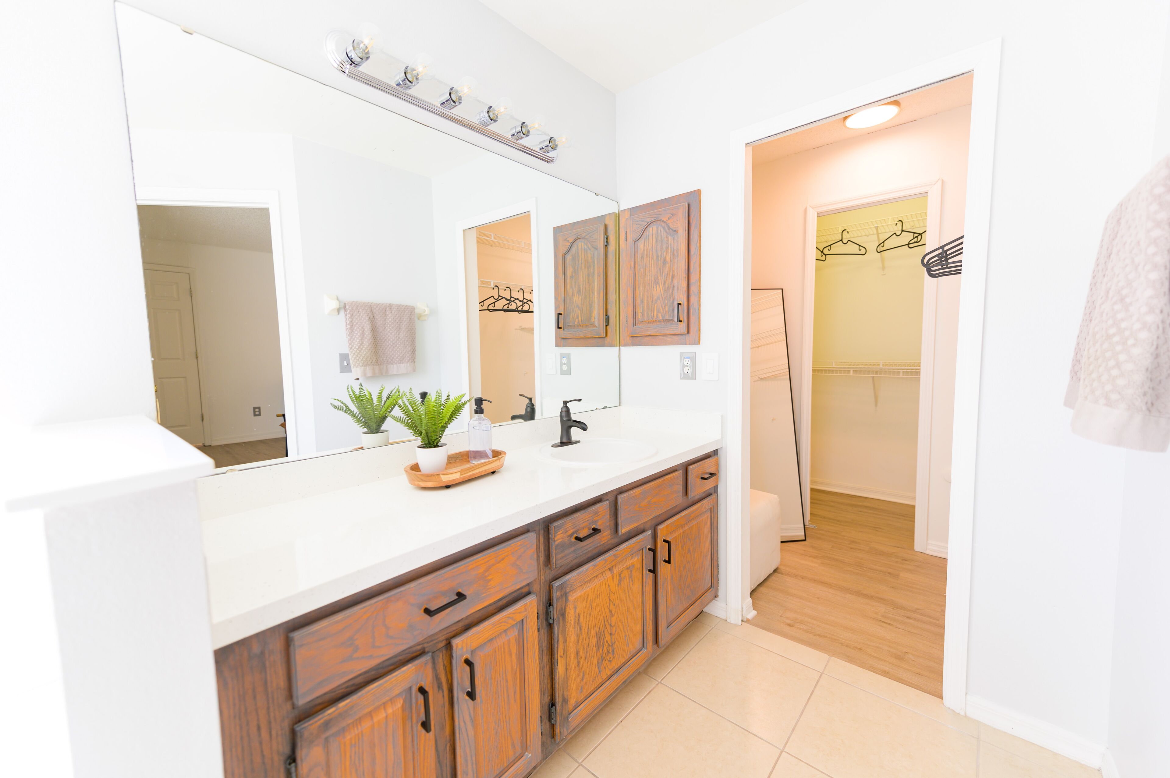 Une salle de bain moderne avec un grand miroir, des meubles en bois, un lavabo agrémenté d’une plante et un dressing avec un portant à vêtements.
