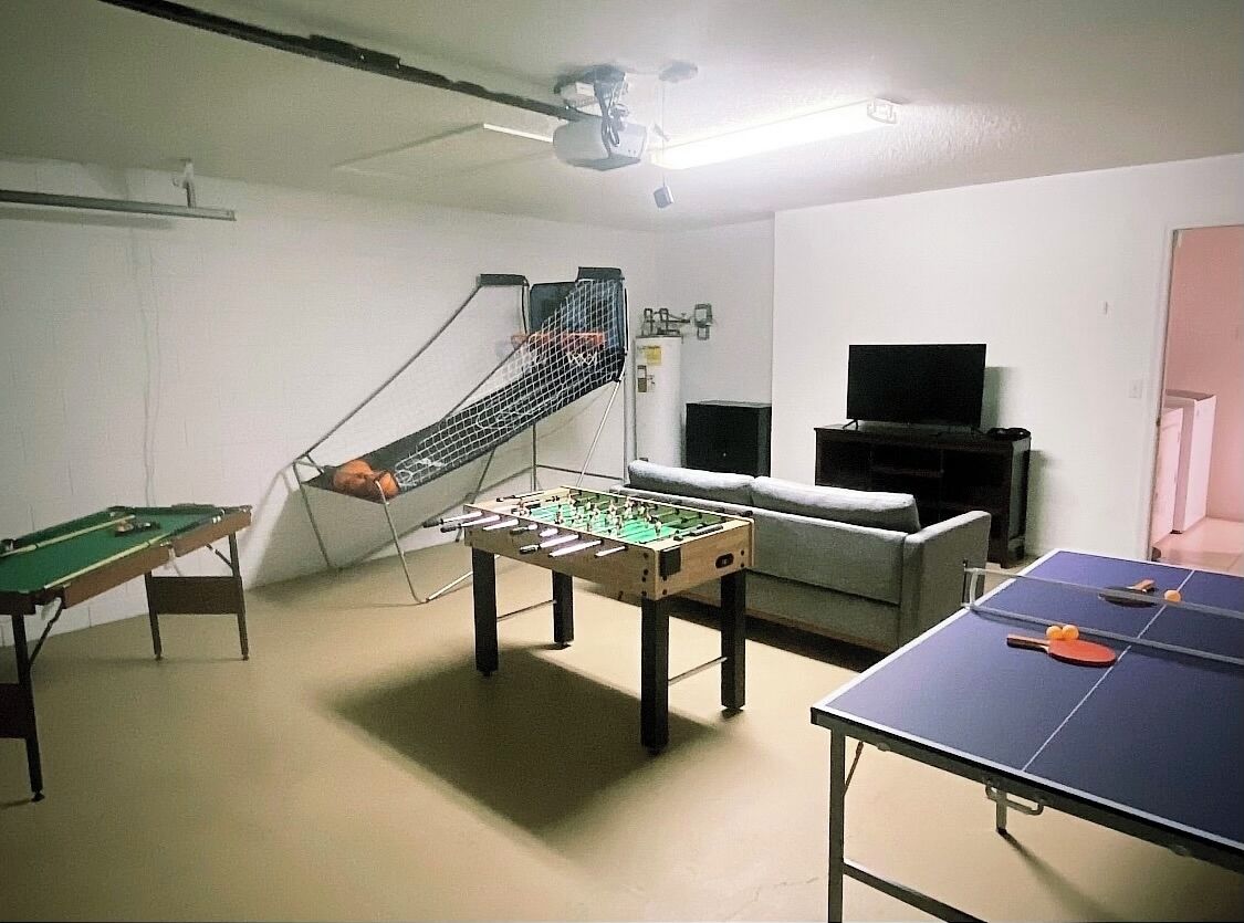 A room with a foosball table, a ping pong table, a pool table, a sofa, and a television.
