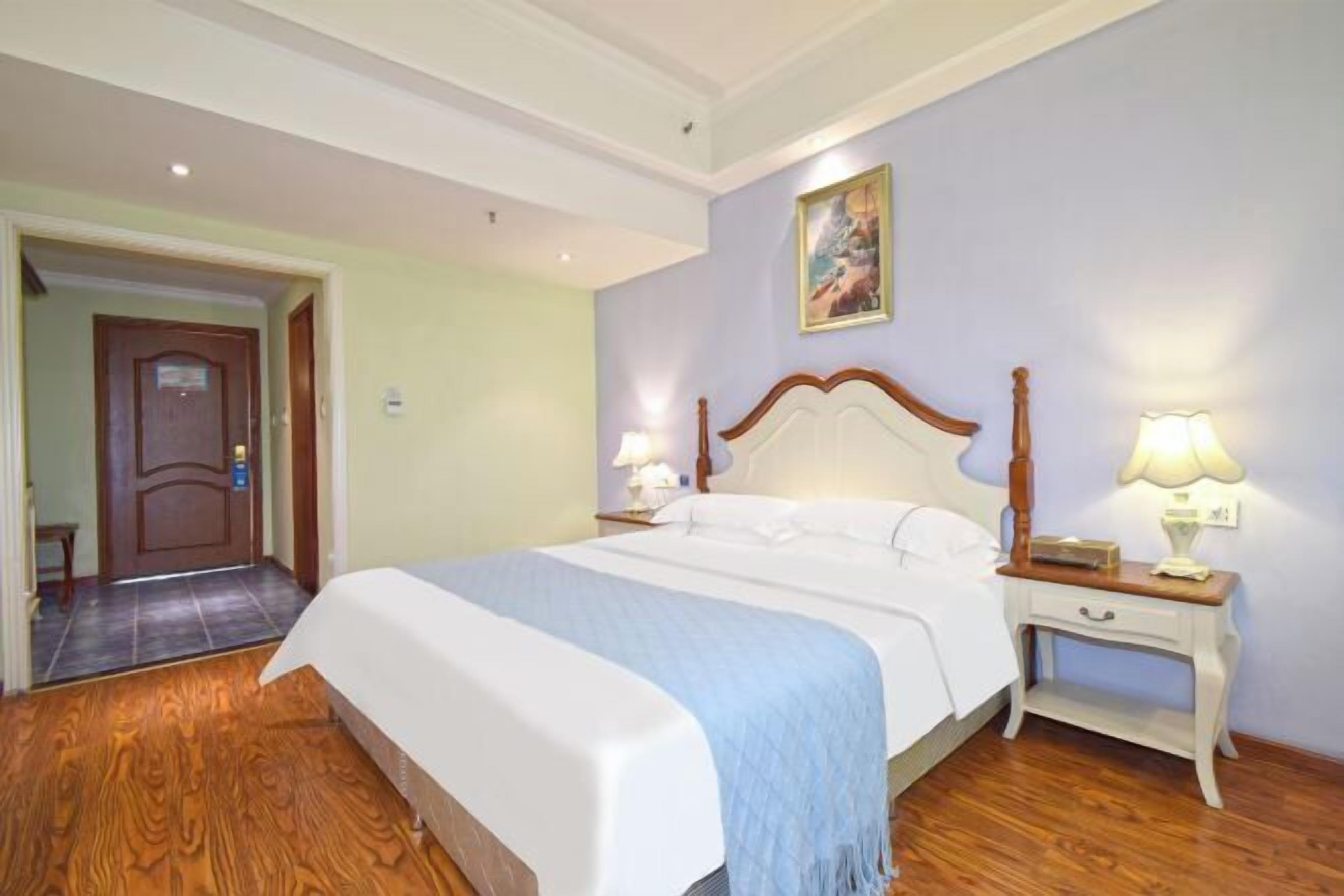 Una habitación de hotel con una cama grande, piso de madera, una puerta y un cuadro en la pared.