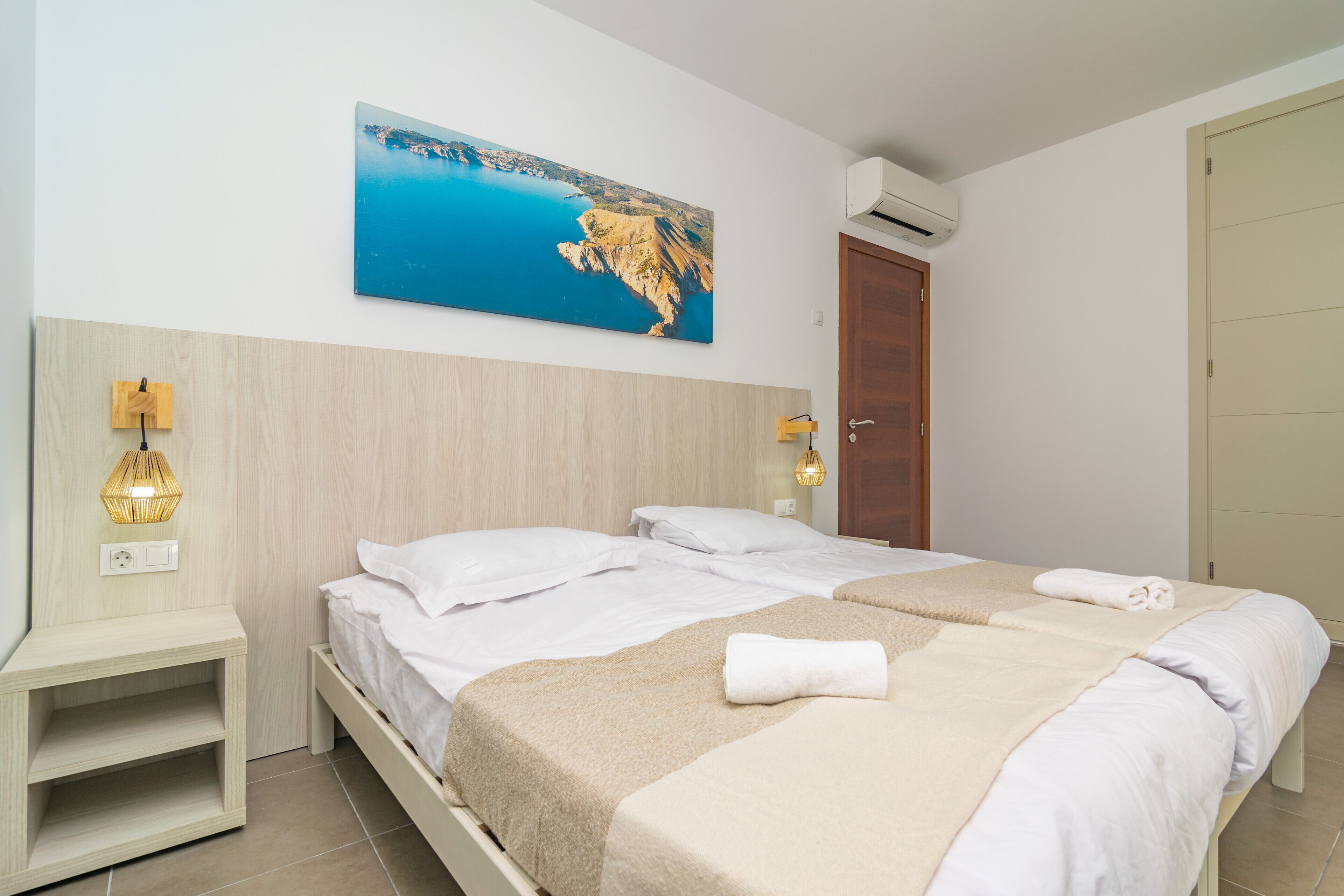 Ein Hotelzimmer mit zwei Betten, einem Gemälde einer Küstenlandschaft, einem Holz-Kopfteil, einem Nachttisch mit Lampe und einer Klimaanlage.