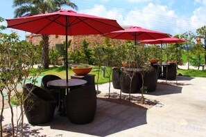 Outdoor seating with wicker chairs and red umbrellas, a concrete patio, and a landscaped garden with palm trees.