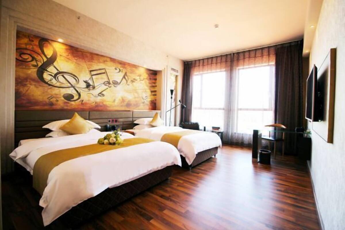 A hotel room with two beds, a large wall mural with musical notes, a large window with curtains, and a flat-screen TV mounted on the wall.