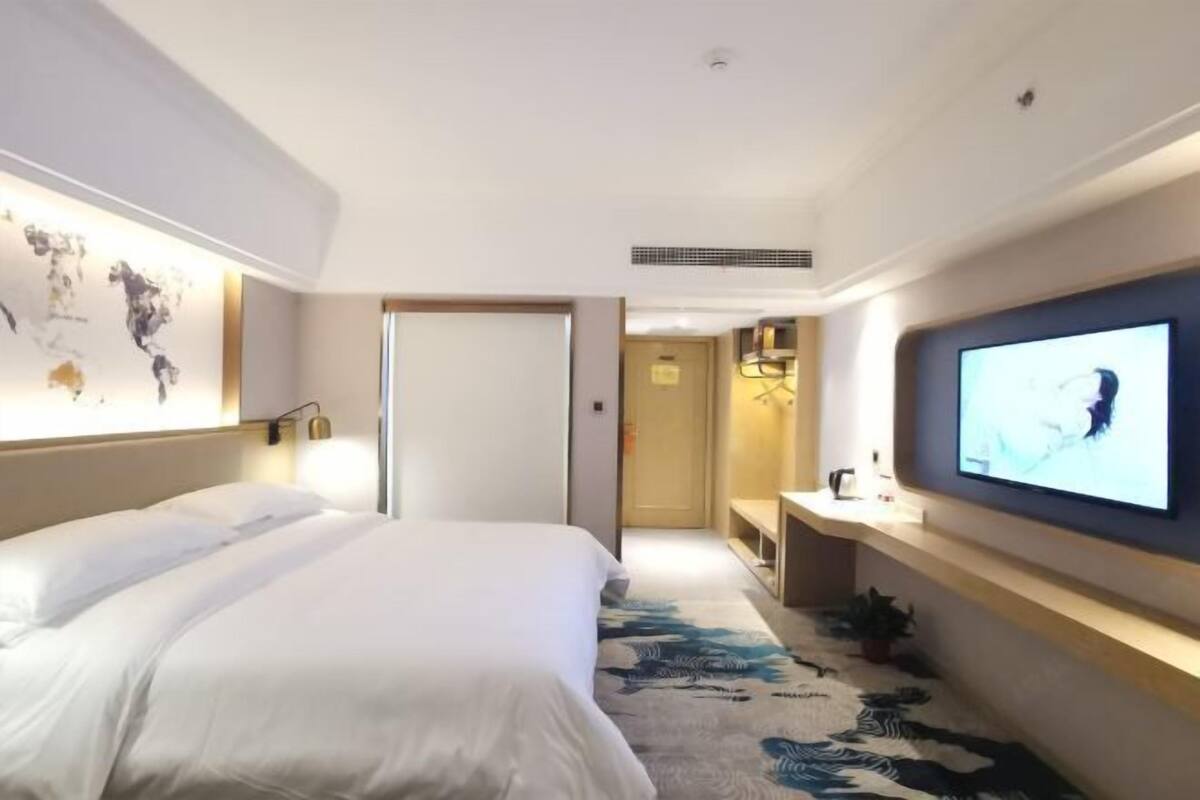 A hotel room with a large bed, a flat-screen TV mounted on the wall, a world map on the wall, and a small desk with a lamp.