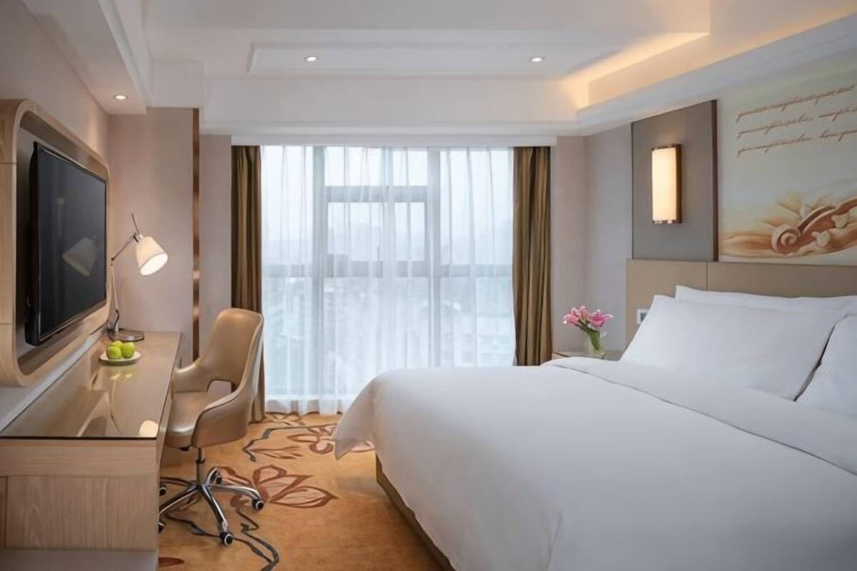 A hotel room with a bed, a desk with a TV, a chair, a desk lamp, a flower vase, and a window with curtains.