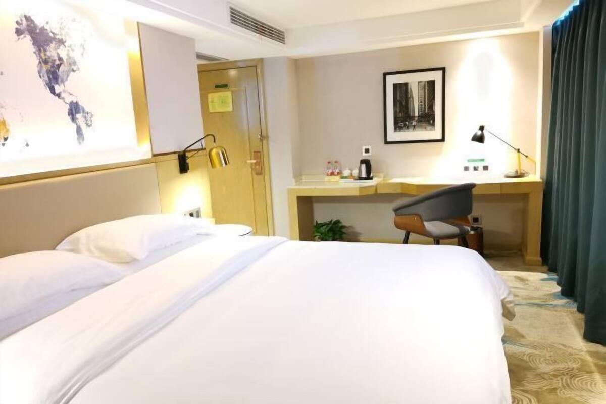 A hotel room with a bed, a desk with a chair, a desk lamp, a framed picture on the wall, and a map on the wall.