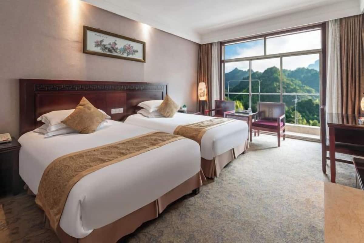 A hotel room with two beds, a desk, a chair, and a large window offering a mountain view.