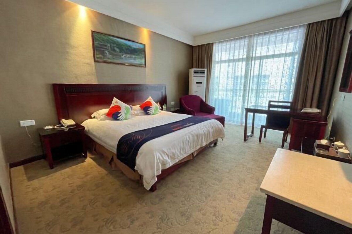 A hotel room with a large bed, a desk, a chair, a telephone, and a window with curtains.