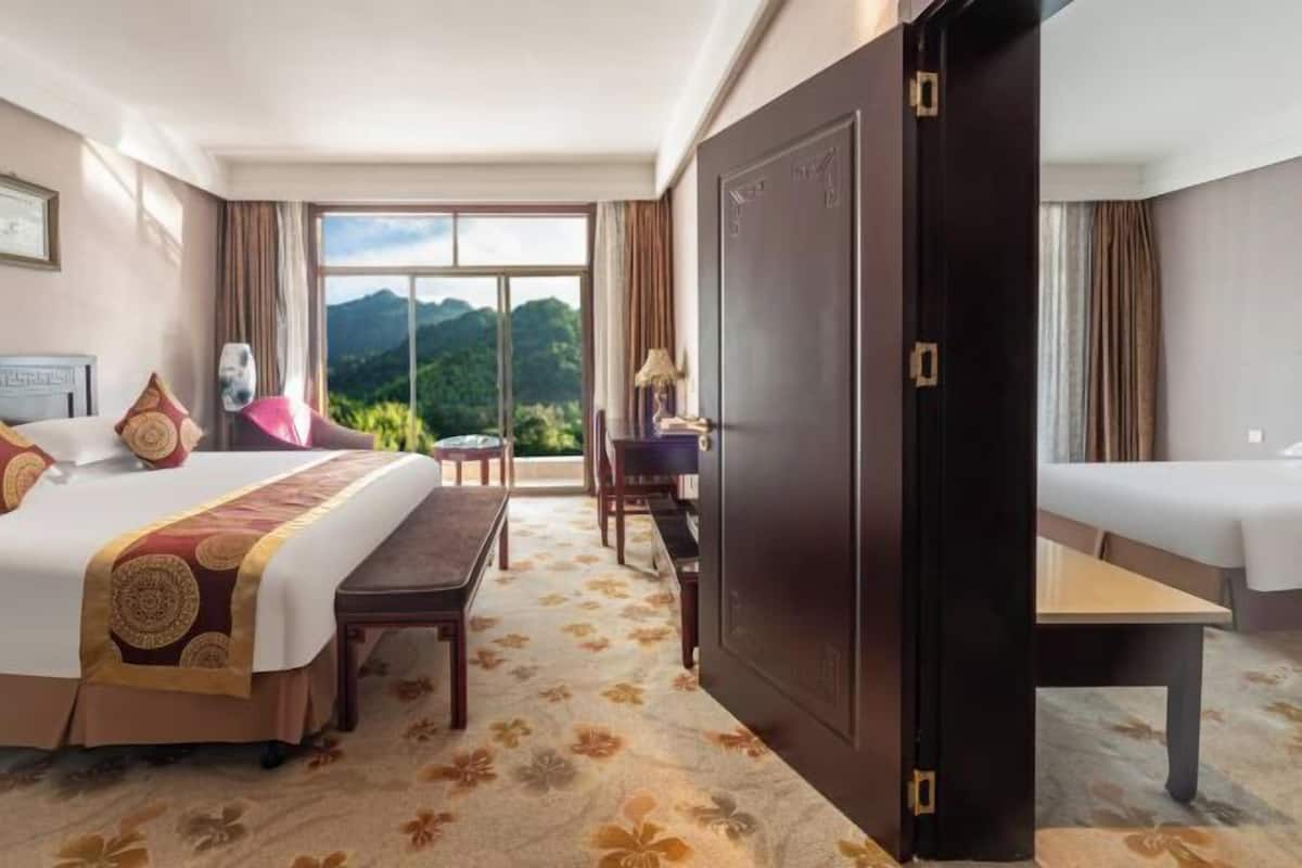 A hotel room with a bed, a desk, a chair, and a view of mountains through a large window.