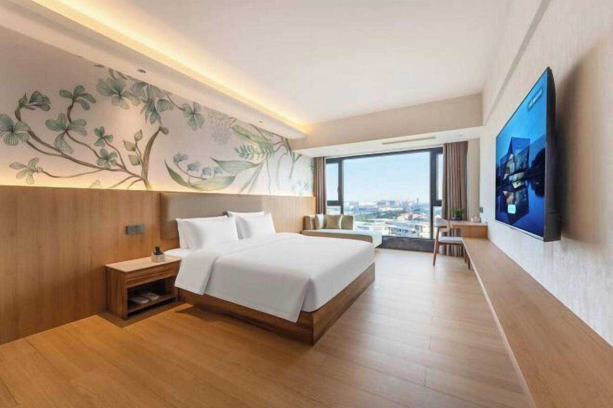 A modern hotel room with a large bed, a desk, a flat-screen TV, and a city view.