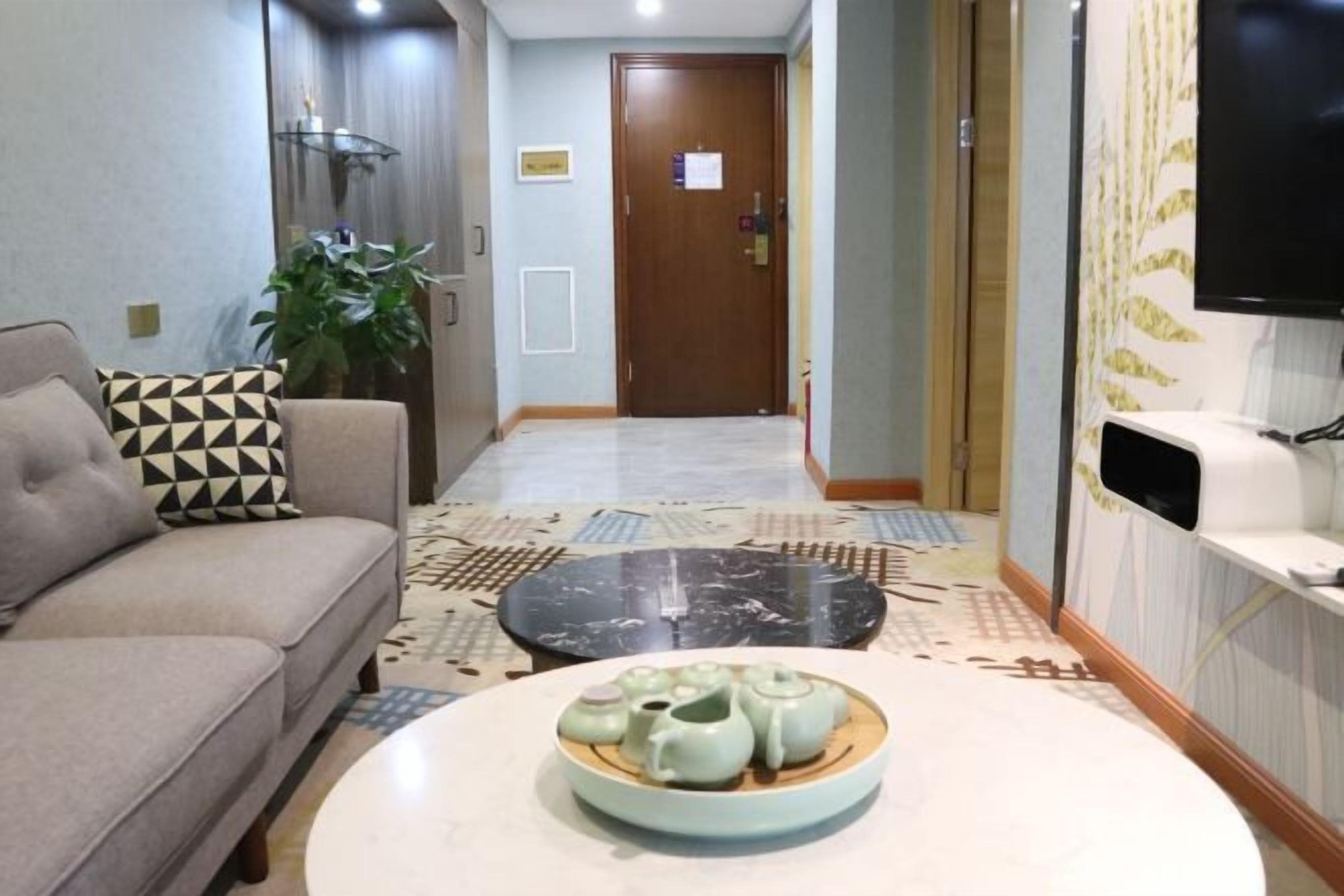A hotel lobby with a sofa, a coffee table with teapots, a plant, a wooden door, and a television on the wall.