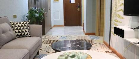 A hotel lobby with a sofa, a coffee table with teapots, a plant, a wooden door, and a television on the wall.