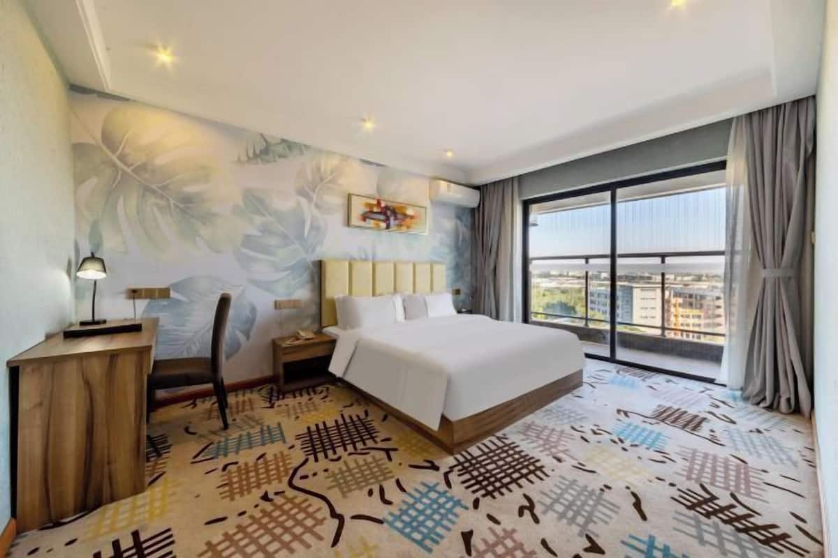 A hotel room with a bed, a desk, a chair, a nightstand, a large window with a view, and a patterned carpet.
