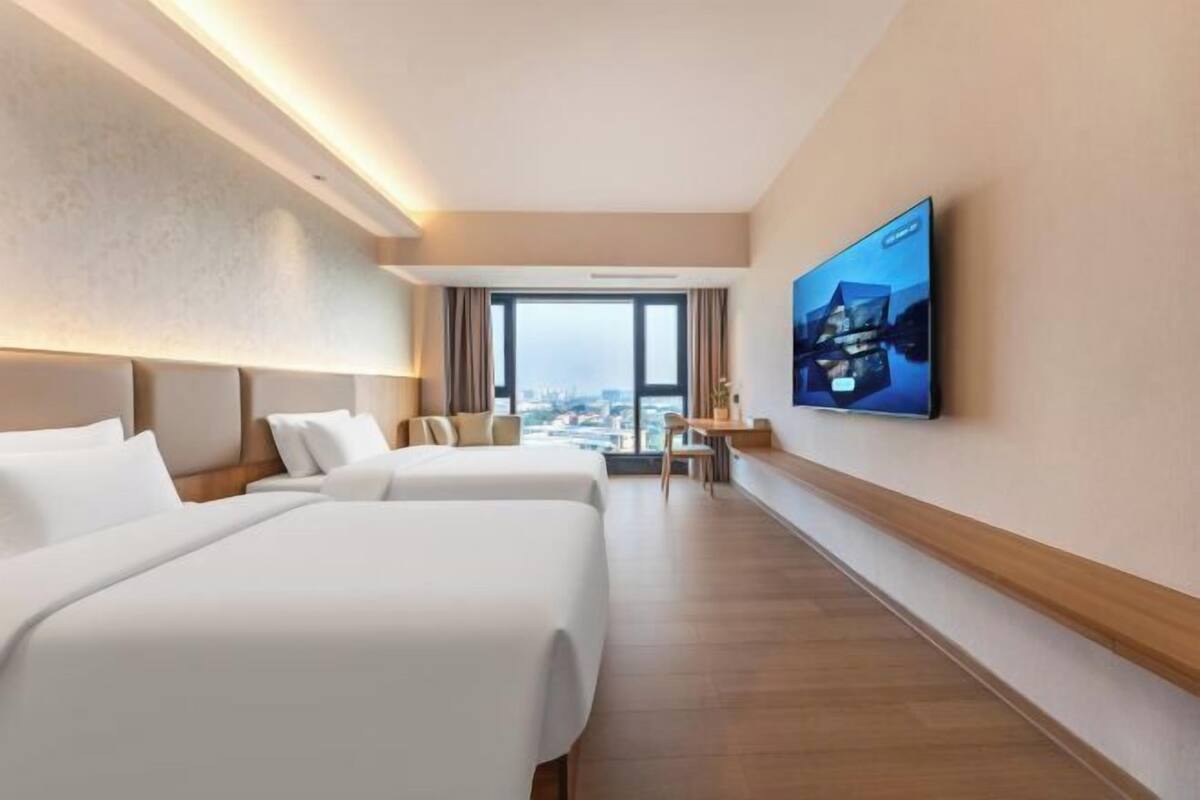 A hotel room with two beds, a TV on the wall, a desk, and a view of the city.