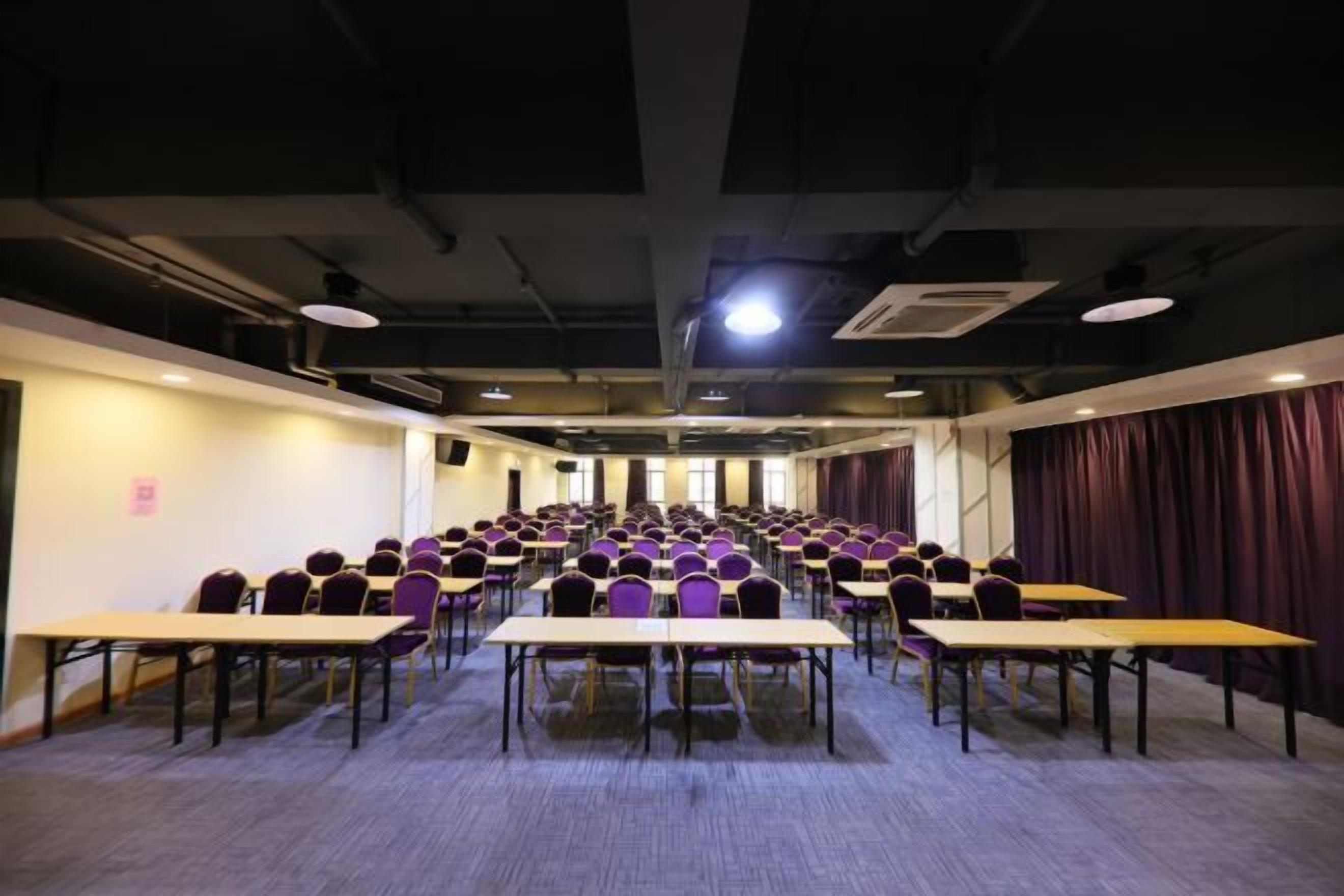 A conference room with rows of tables and chairs, a carpeted floor, and a stage area with curtains.