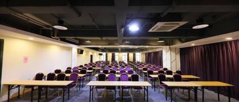 A conference room with rows of tables and chairs, a carpeted floor, and a stage area with curtains.