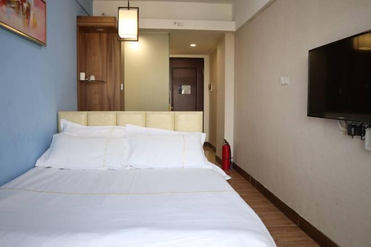 A hotel room with a bed, a television mounted on the wall, a wooden wardrobe, and a door leading to another room.
