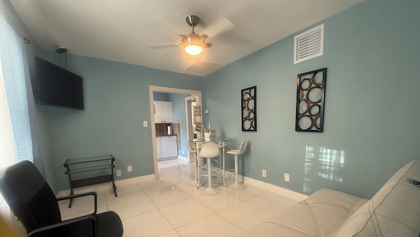 A living room with a white sofa, a black office chair, a dining table with chairs, a ceiling fan, and artwork on the walls. - Cute Paradise 2 (Tampa)