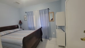 A bedroom with a bed, a nightstand, a lamp, a wardrobe, and a window with curtains. - Cute Paradise 2 (Tampa)