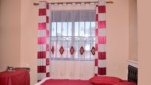 A bedroom with a bed, red bedding, a table with an ironing board, and a window with decorative curtains. - Stunning 2-Bed Apartment in Namugongo Town Kampala (Namugongo Town)