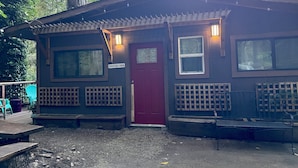 Eine Hütte mit roter Tür, zwei Fenstern, einem Schild mit der Aufschrift 'CREEKSIDE CABIN' und einem Holzzaun.