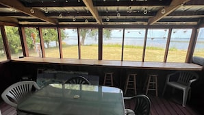 Une terrasse extérieure couverte, dotée d’une table et de chaises en verre, de poutres en bois et offrant une vue sur une étendue d’eau.