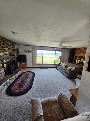 A spacious living room with a stone fireplace, a large window offering a view of the outdoors, a ceiling fan, and a cozy seating area. - Home with panoramic lake views of Lake Poygan  (Winneconne)