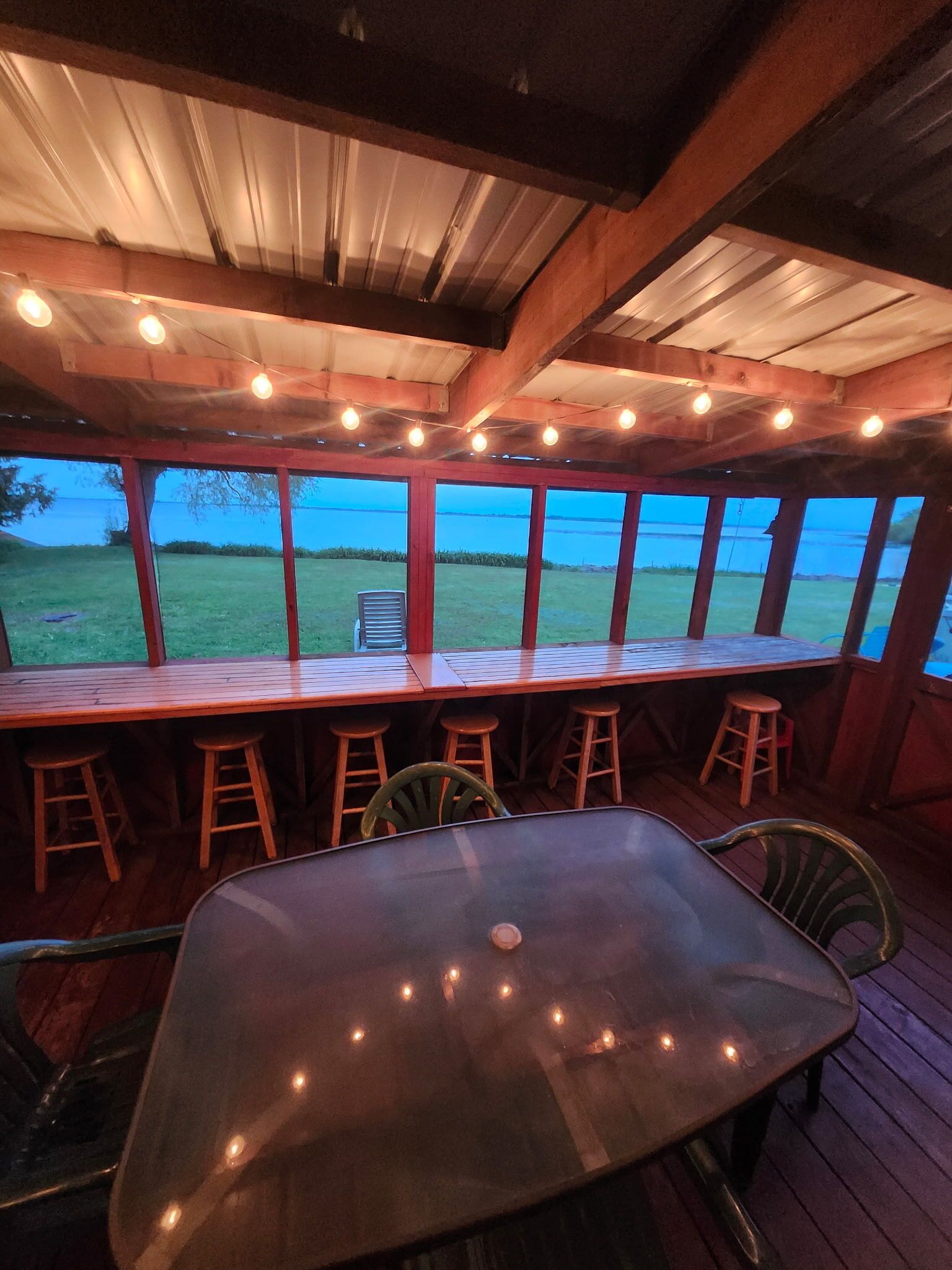 A covered outdoor seating area with a table and chairs, overlooking a grassy field.