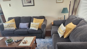 Ein Wohnzimmer mit zwei grauen Sofas, jeweils mit einem gelben Kissen und einem 'relax'-Kissen. Vor den Sofas steht ein Holztisch mit einer Schale grüner Äpfel und einer Fernbedienung.