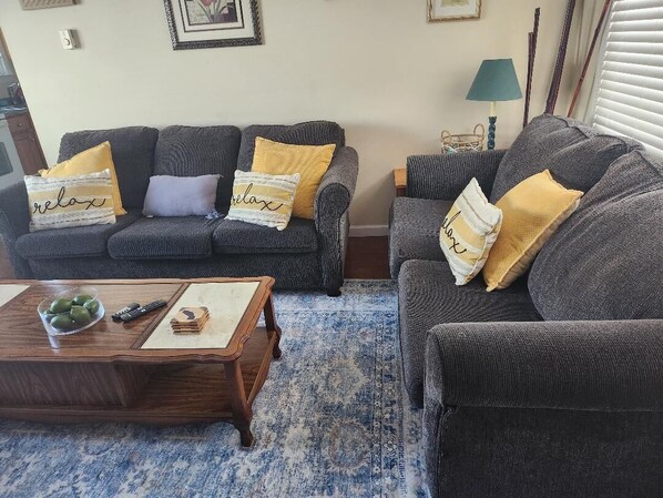 A living room with two grey sofas, each with a yellow pillow and a 'relax' pillow. A wooden coffee table with a bowl of green apples and a remote control is in front of the sofas.