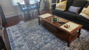 Ein Wohnzimmer mit einem dunkelgrauen Sofa, einem Holztisch mit einer Obstschale und einem Bücherstapel, einem gemusterten Teppich sowie einem Essbereich mit Tisch und Stühlen.