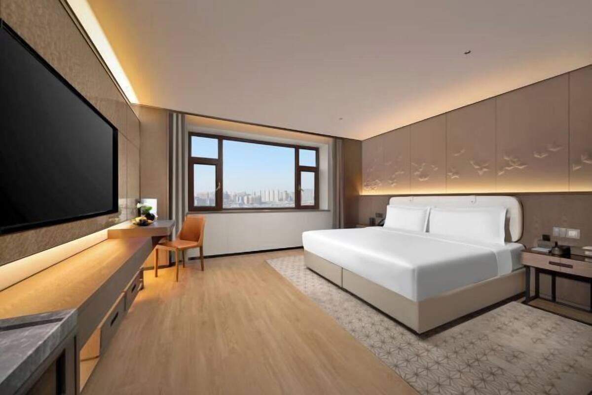 Ein modernes Hotelzimmer mit einem großen Bett, einem Schreibtisch, einem Sessel, einem Fernseher und einem großen Fenster mit Blick auf die Stadt.