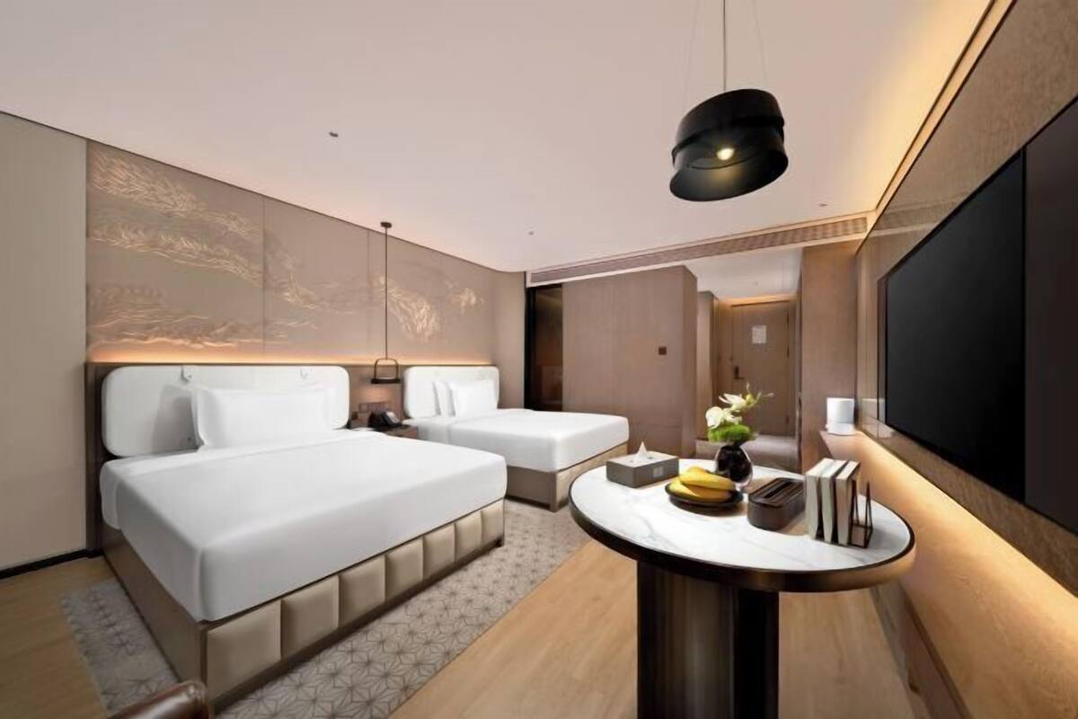 Ein modernes Hotelzimmer mit einem großen Bett, einem Einzelbett, einem runden Tisch mit einer Vase und Büchern sowie einem großen Fernsehbildschirm.