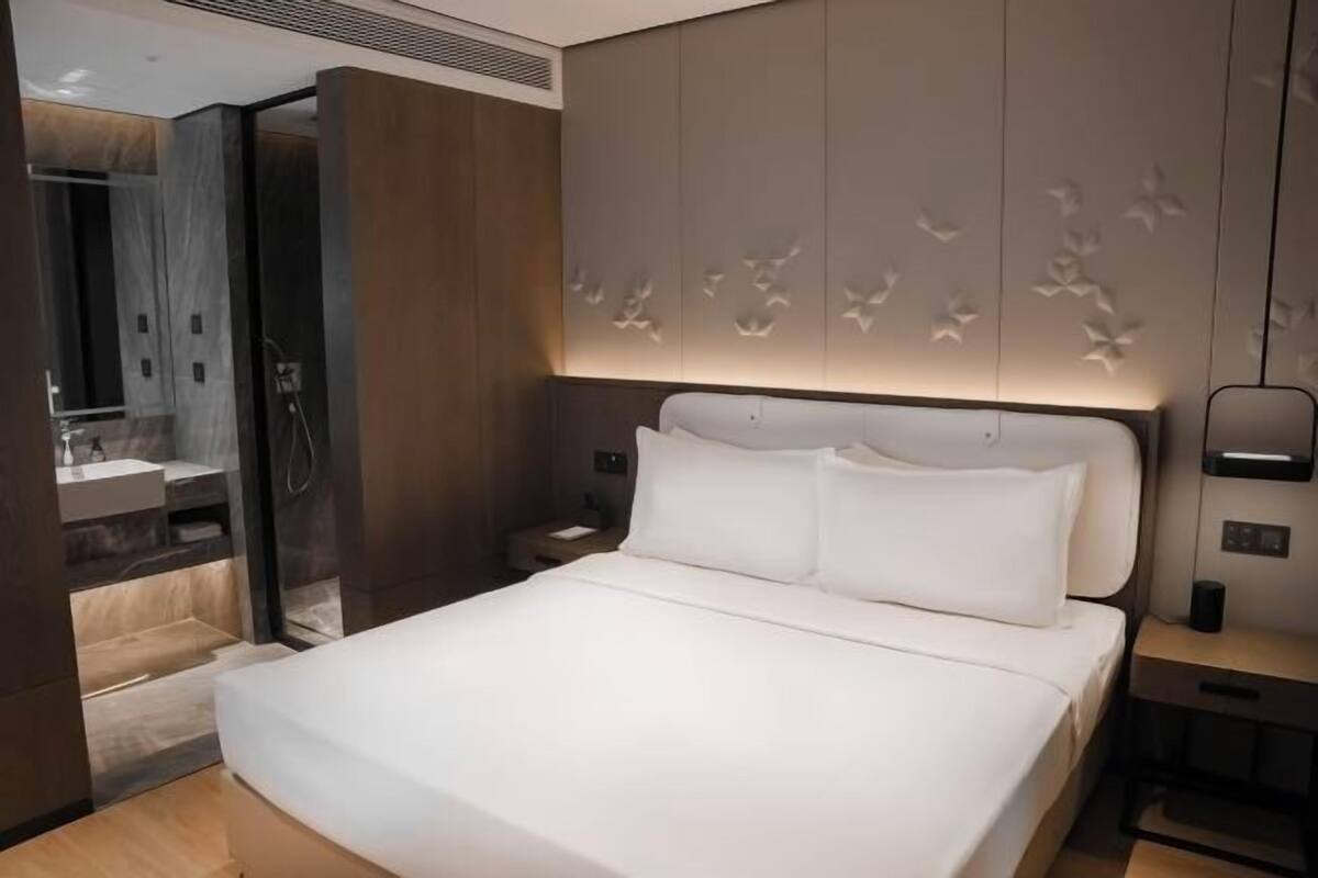 Ein modernes Hotelzimmer mit einem großen Bett, einem Nachttisch, einer an der Wand befestigten Lampe und einem durch eine offene Tür einsehbaren Badezimmer.