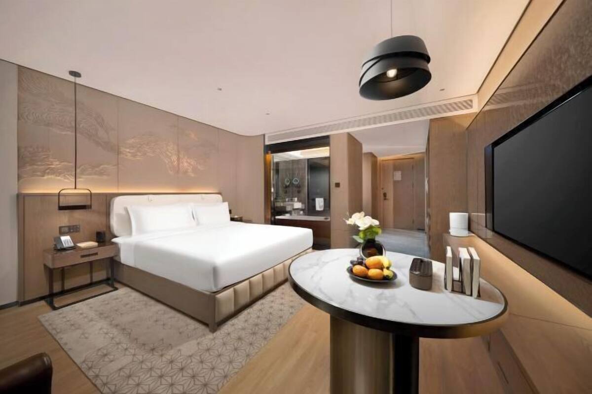 Ein modernes Hotelzimmer mit Bett, Nachttisch, Lampe, Fernseher und einem kleinen runden Tisch mit Obst und Büchern.