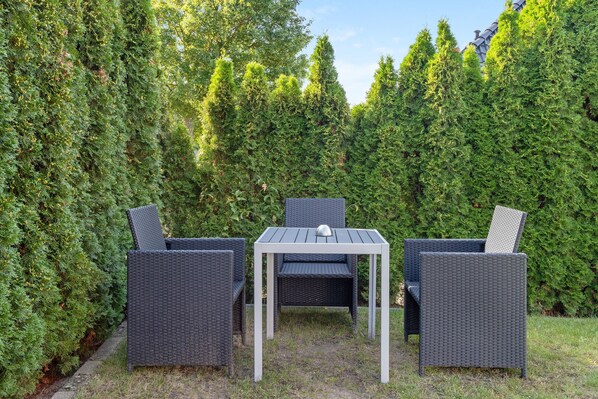 Ein Patio mit Korbmöbeln, darunter ein Tisch und zwei Stühle, umgeben von dichtem Grün.