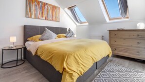 Une chambre à coucher comprenant un lit, une table de chevet, une commode, un puits de lumière et un tableau accroché au mur.