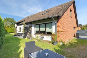 Une maison avec un toit en pente, une façade en briques et un petit jardin devant, avec une table et des chaises de patio.