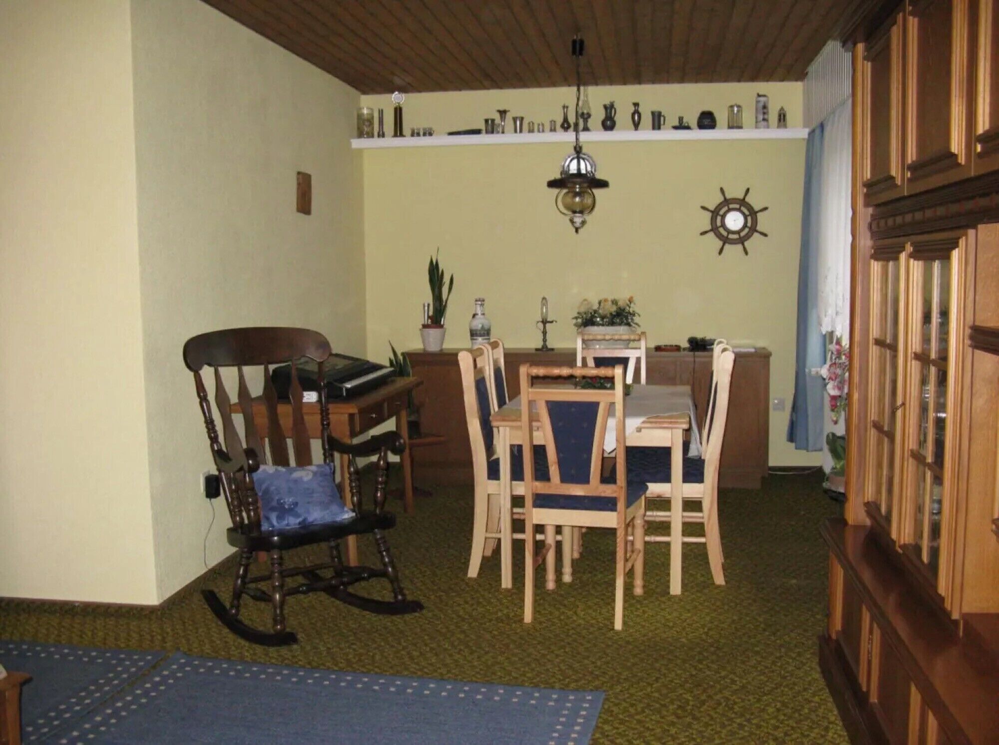 A dining area with wooden chairs, a table, a rocking chair, a chandelier, and a wall clock.