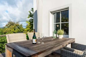 Une table en bois avec une bouteille et des verres, des chaises en osier, une plante en pot et une fenêtre décorée d’un motif en forme de cœur.