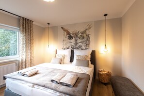 Een slaapkamer met een bed, kussens, een kat op wanddecoratie, nachtlampen en een raam met gordijnen.